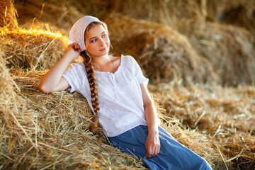 Beautiful blonde girl with braided hair in white rural clothes sitting on a straw. Farm life.Young model posing on a ranch.Warm art work.