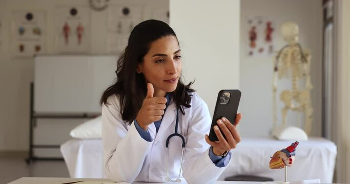 Attractive Latina female cardiologist in uniform calling to patient use video conference application on cellphone, provide professional help remotely, talking to colleague take part in medical council