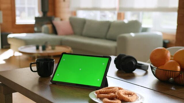 Tablet computer with green screen chromakey display with motion trackers lay on table near oranges, cookies and cup of hot tea and sofa on background.cozy interior of living room in house on daylight