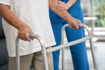 Female patient holding a walker and trying to practice walking in hospital. Nurse helps her walk at...