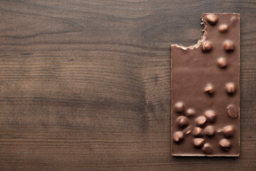 Fototapeta na wymiar Bitten chocolate bar with whole hazelnuts on brown table. Flat lay image of chocolate on wooden background with copy space.