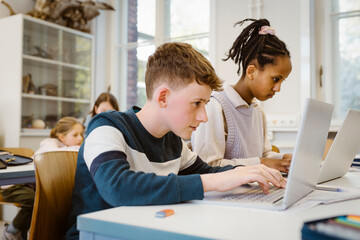 Focused schoolboy using laptop by female friend at desk in classroom
