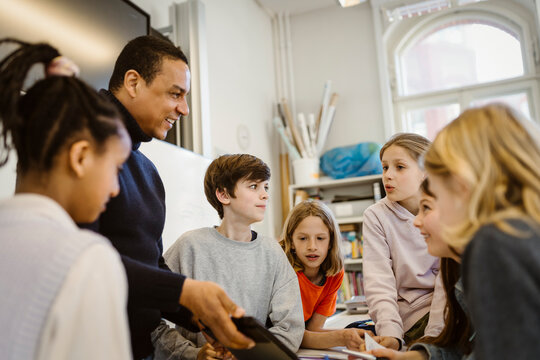 Male teacher sharing tablet PC with students discussing in classroom