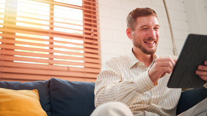 Caucasian man, Bearded smiling using touch screen digital tablet. While sitting on sofa in living room at home.