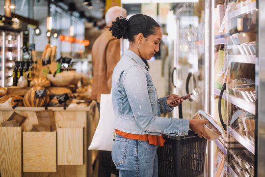 Side view of female customer photographing package while standing at grocery store