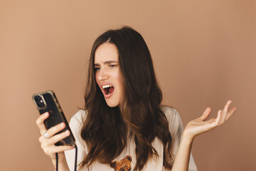 Mad and shock young brunette beautiful woman using mobile phone isolated on beige background. Yelling unhappy woman hold mobile phone. Disappointed sad upset lady horrified impressed news. WTF. Oh no.