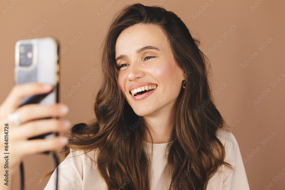 Wall mural Young smiling brunette curly woman wear beige t-shirt doing selfie shot on mobile phone post photo on social network isolated on beige background. Woman make video call, selfie on her mobile phone.