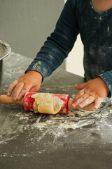 Cute toddler boy baking Christmas cookies at home. Red rolling pin and cookie cutters on dark grey kitchen table. 