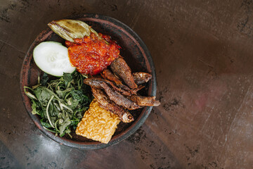 Flat lay shot of a wooden plate of Indonesian traditional food called Lalapan contains fried Iwak Wader or Spotted Barb fish, vegetables, tempe, cucumber and eggplant with sambal. Selective focus.