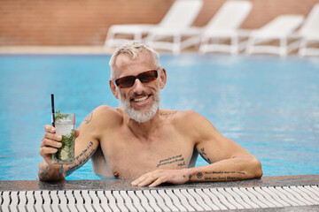 shirtless and tattooed middle aged man in sunglasses holding cocktail and swimming in pool, retreat