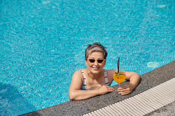 positive mature woman in sunglasses swimming in pool with blue water and holding cocktail, retreat