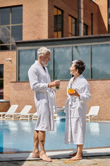 cheerful middle aged couple in white robes holding cocktails at poolside, wellness retreat in resort