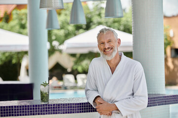 cheerful mature man in white robe standing at pool bar near glass with mojito cocktail, retreat