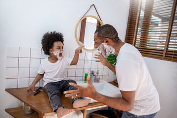 Portrait of happy  African American black father son apply shaving foam on face while looking in reflection mirror morning routine. Man shaving with razor, hygiene body care, family lifestyle concept