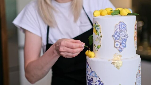 Satisfied housewife putting decorative green leaf near yellow lemons on top of two-tiered cake standing in kitchen small business of organic sugar free confectionery production at home