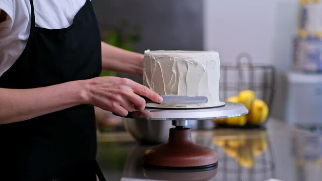 Skilled confectioner smearing sugar free white cream around baked cake with knife on blurred background woman in black apron creating delicious custom pastry for events closeup
