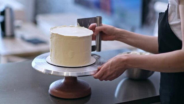 Housewife in black apron processing white mastic on organic lactose free cake with pastry scraper standing at table woman using kitchen utensils to make delicious confectionery at home
