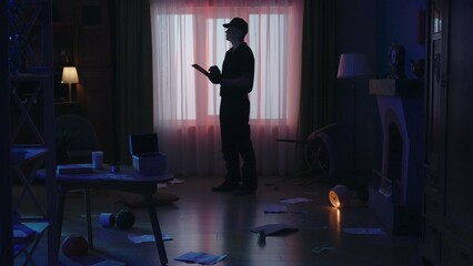 Crime scene creative concept. Man officer with tablet standing in the dark living room. Flashlights behind the window.
