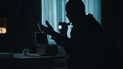 A thief in a black balaclava commits a burglary, stealing valuables. Criminal in a dark apartment...
