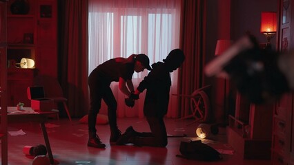 The arrest of the robber in a dark apartment, lit with red light. Side view of a criminal kneeling at gunpoint with his hands behind his back. A female police officer handcuffs the villain.