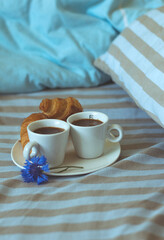 Two cups of espersso and a croissant in bed on a spread out bed.