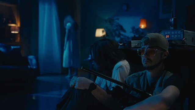 Two ghost hunters hiding behind the table. A female silhouette , ghost, entity standing on the background. The men are talking, scared and frightened.