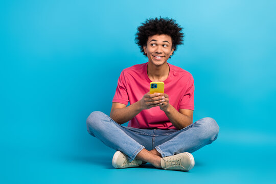 Full length photo of nice young guy sit floor look empty space gold device dressed stylish pink garment isolated on blue color background