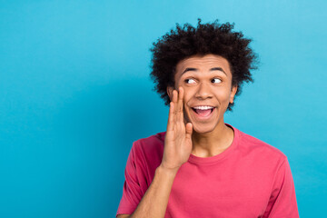 Portrait of funny guy with afro hairdo dressed stylish t-shirt hand on cheek tell secret to empty space isolated on blue color background