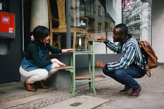 Female upcycling store owner showing furniture to customer while crouching at sidewalk