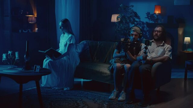 Video of two ghost hunters sitting on the side of the couch trying to capture on video a posessed female, ghost, poltergeist sitting in front and reading.