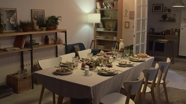 Festive table in rectangular form with white tablecloth with dishes and wine on it, chairs around it in living room with cozy interior in pastel colors