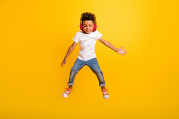 Fototapeta na wymiar Full length photo of positive cheerful little boy dressed white t-shirt jumping high enjoying songs earphones isolated yellow color background
