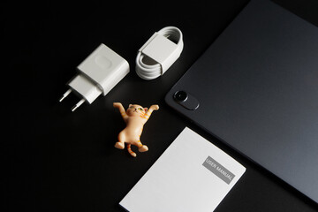 Modern gray electronic tablet with a metal case lies on a black background next to a charger - power supply, usb cable, user manual and a toy cat. Gadget unboxing. Photo