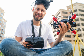 Outdoor portrait of young smiling professional fpv drone pilot wering goggles and posing with remote controller and copter in hands