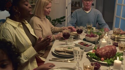 Members of multi-ethnic family praying before festive dinner sitting by served table with dishes and wine in cozy living room