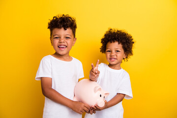 Photo of friendly cheerful little kids hold money bank pig demonstrate v-sign isolated on yellow color background