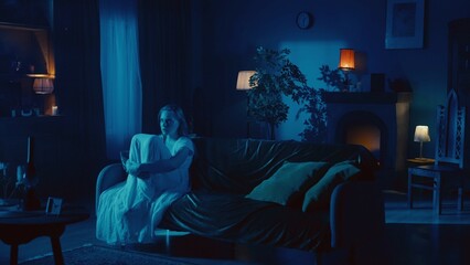 Shot of a young woman sitting on the couch, embracing, hugging her legs and mumbling, whispering something as if she is posessed by a ghost, demon, poltergeist. - obrazy, fototapety, plakaty