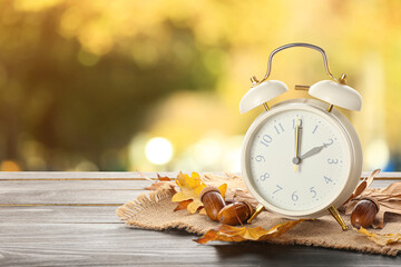 Alarm clock, acorns and autumn leaves on table outdoors. Daylight saving time end - obrazy, fototapety, plakaty