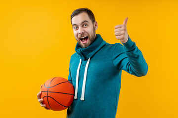 Studio shot of young attractive basketball fan posing over bright colored orange yellow background holding the ball in hand and showing thumbs up gesture