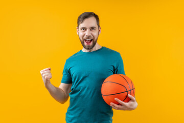 Studio shot of young attractive basketball fan posing over bright colored orange yellow background holding the ball in hand and making winner's gesture clenching his fist