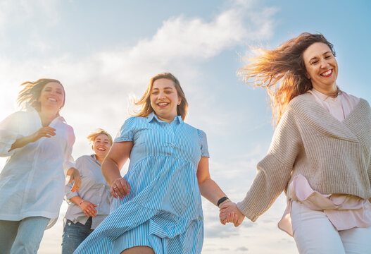 Portrait of four cheerful smiling women holding hand in hand running by the meadow. Low angle photo shot on the blue sky with clouds. Woman's friendship, relations, and happiness concept image.