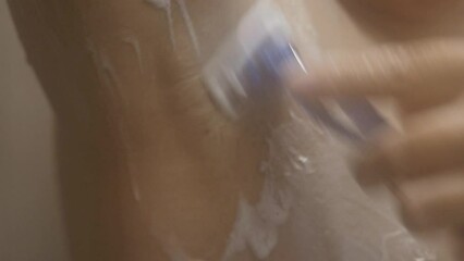 A woman stands in the shower, shaves her hair with a razor under her arm, raising her hand up. Around steam from the shower, selective sharpness, close-up.