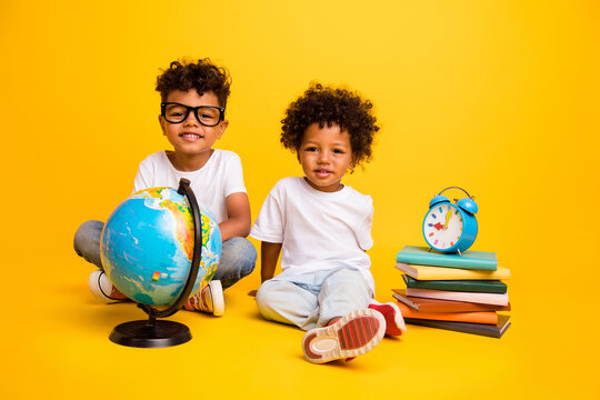 Full size photo of cheerful clever schoolkids sit floor pile stack book world globe bell ring clock isolated on yellow color background