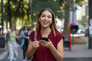 Happy beautiful young woman looking at camera and holding mobile phone outdoor with blurred people...