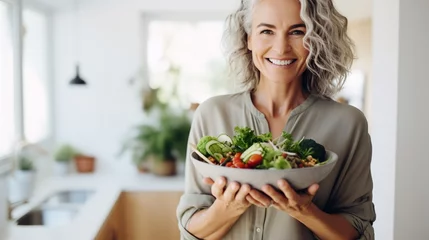 Fotobehang Aged woman smiling happily and holding a healthy vegetable salad bowl on blurred kitchen background, with copy space. © JW Studio