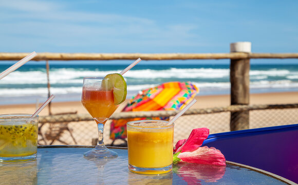 Drinks tropicais em mesa de praia no nordeste do Brasil. No fundo ondas do mar e um guarda sol colorido