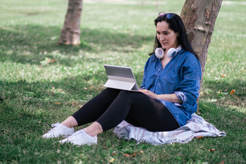 Young woman with tablet computer and cordless earphones sitting on lawn and leaning on tree trunk in city garden remote employee working on internet in park