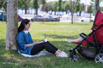 Young woman with headphones working on tablet computer while baby sleeping in buggy on summer park lawn mother surfing internet via device walking with infant child