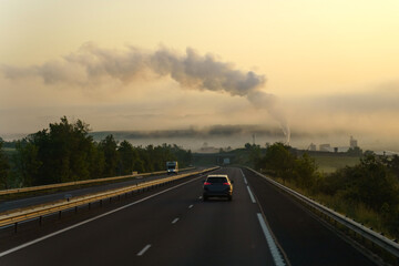 Near the motorway there is an industrial enterprise from the chimney of which there is thick smoke, in the rays of the rising sun