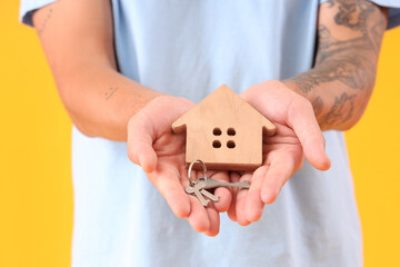Young man with keys and house figure on yellow background, closeup